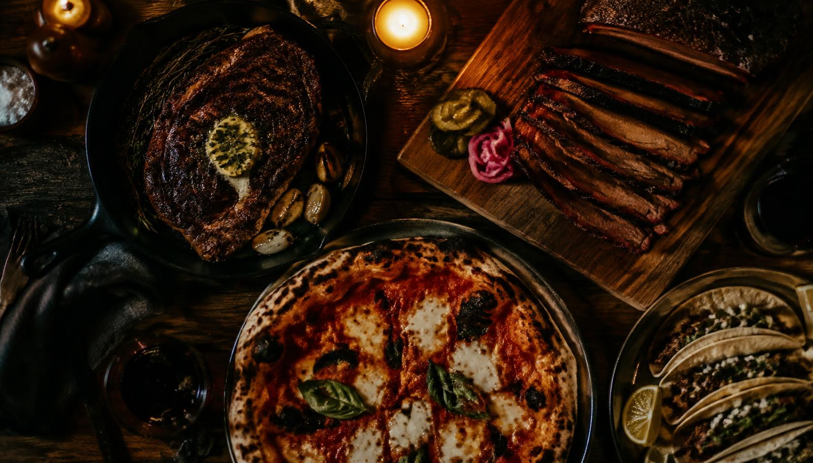 Steak, brisket, pizza and tacos on a dark wooden table